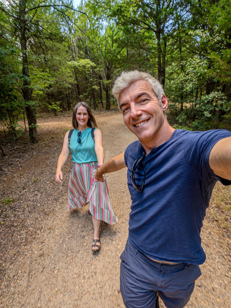 A creator taking a selfie on a wooded trail as a friend walks up behind him