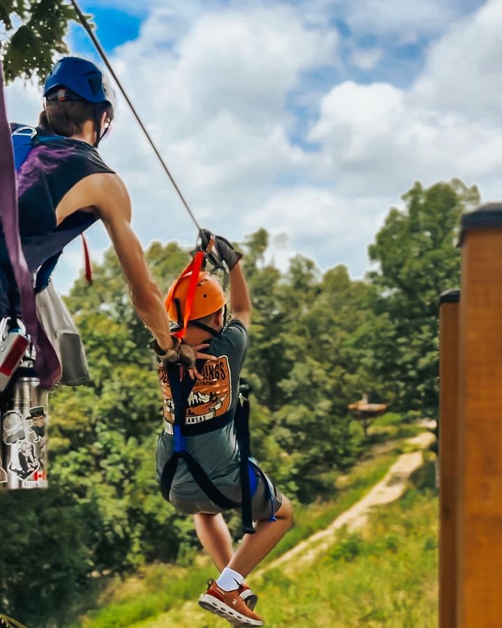 A child in a harness riding a zip line over a wooded hillside