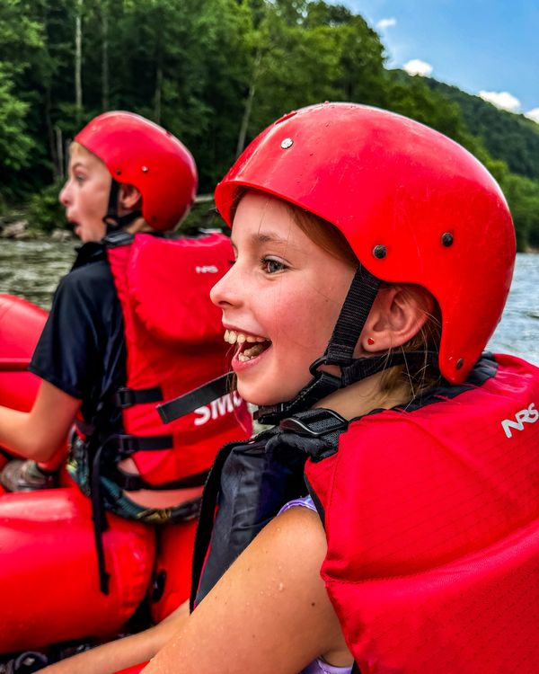 Two kids in red helmets and life jackets laughing on a whitewater raft