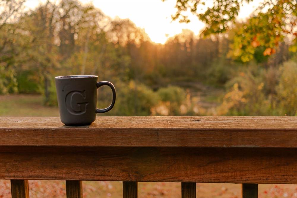 A coffee mug on a cabin deck rail at sunrise, with fall trees beyond